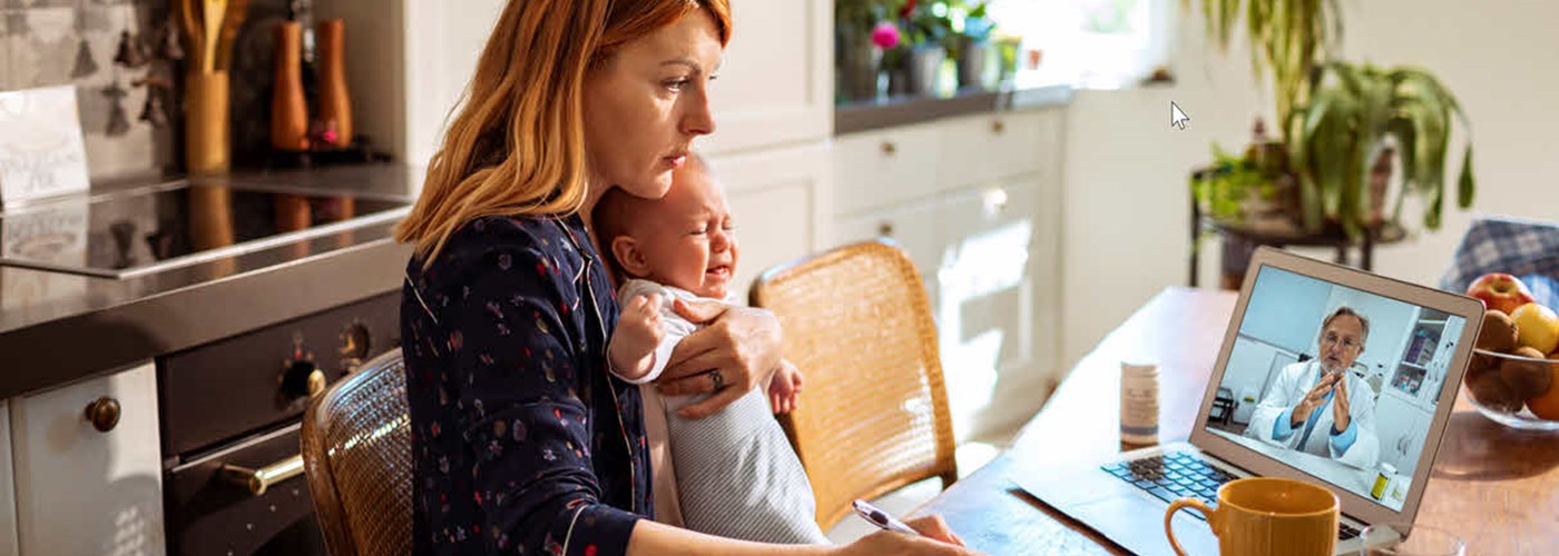 A young mother sat at her kitchen table with her baby. She is speaking to her GP remotely on her laptop.