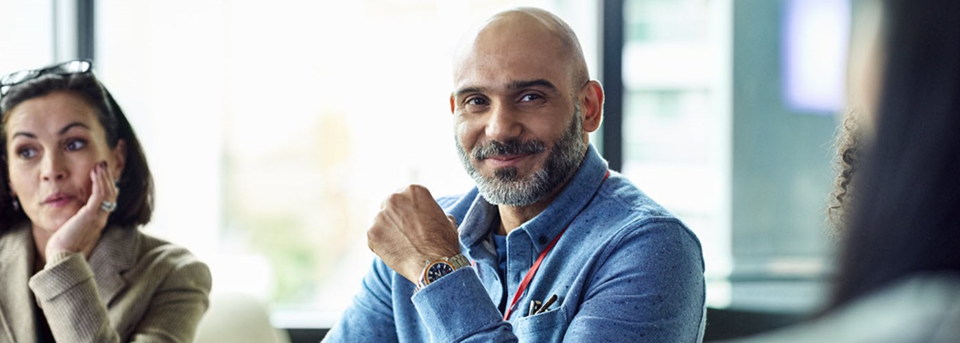 A female and male colleague sat in front of a large window, at a table, having a meeting - listening and smiling 