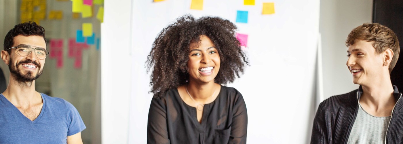 Two gentlemen and one lady smiling at work, in front of a whiteboard covered in post-it notes 
