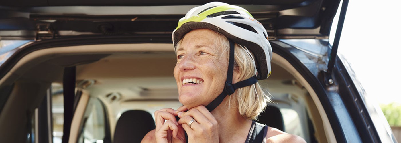 Middle aged woman with short blonde hair in front of her car fastening her cycle helmet whilst smiling 