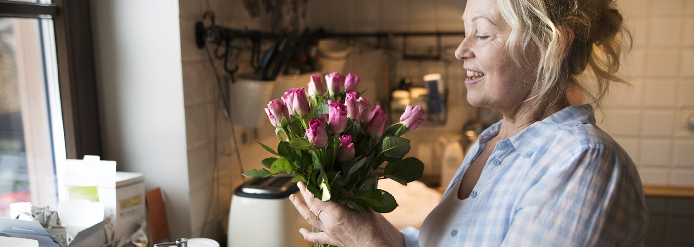 A blonde, smiling middle-aged woman is in her kitchen, arranging fresh flowers.